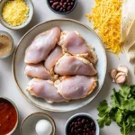 A flat lay of ingredients for slow cooker chicken enchiladas, including raw chicken, black beans, enchilada sauce, cheese, tortillas, spices, and fresh herbs