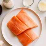 Fresh salmon fillets with measured ingredients including heavy cream, chicken broth, olive oil, garlic, parmesan cheese, sun-dried tomatoes, and spices
