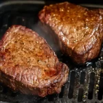 Two juicy, golden-brown steaks resting inside an air fryer basket, showcasing a perfectly cooked crust and flavorful seasoning.